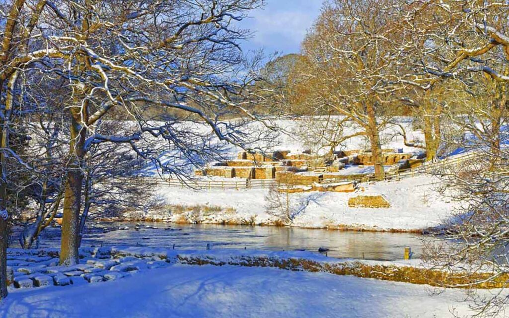 visiting hadrian's wall: a guide to every season. Winter image by Roger Clegg. A view across an icy river to the Roman baths at Chesters. All is covered in snow but the stones of the buildings can still be seen.  