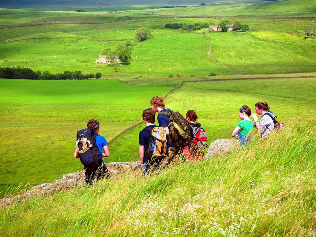 visiting hadrian's wall: a guide to every season. Summer image by Roger Clegg.  A group of six young people have their backs to the camera. Lush, green fields stretch out below them with two sandstone farmhouses on the holls opposite as they rise. Hundreds of sheep can be seen grazing in the distance. 