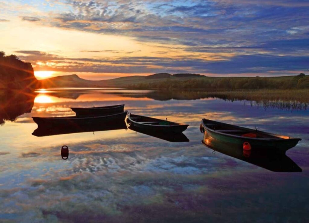 Hadrian's Wall Guide. Three small row boats sit on a lake as the sun sets behind..