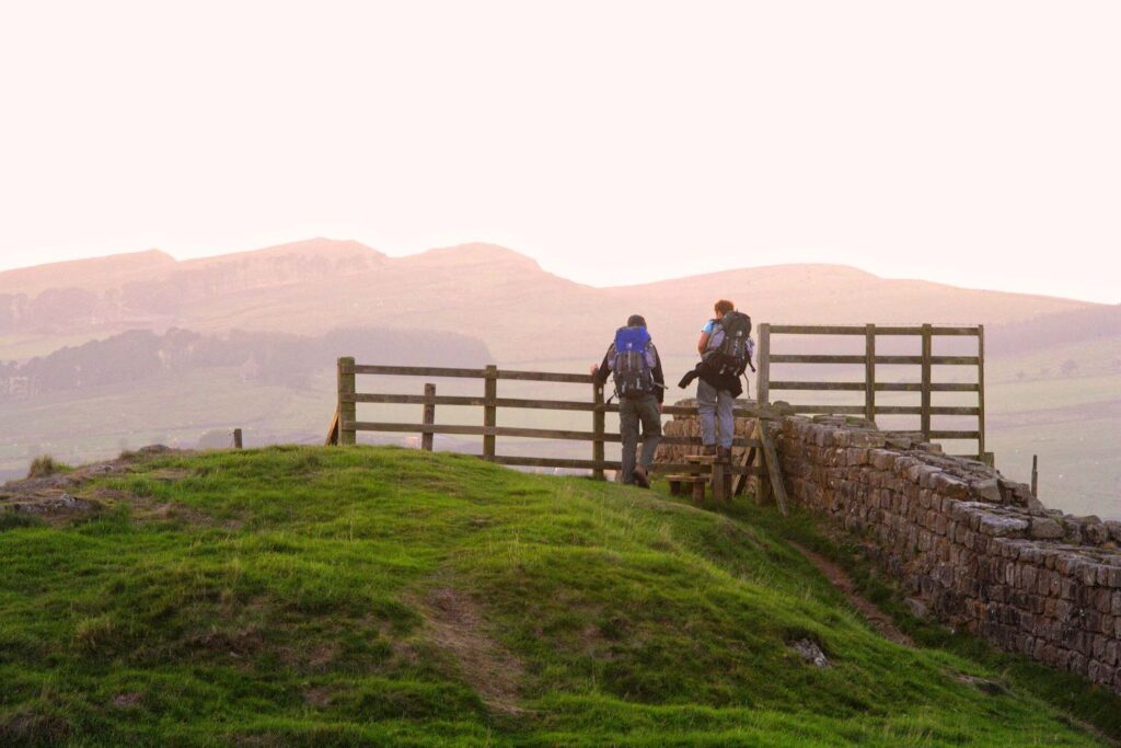 Hadrian's Wall Guide.Two young men wearing backpacks are climbing over a wooden stile with rolling hills and some crags in front of them.