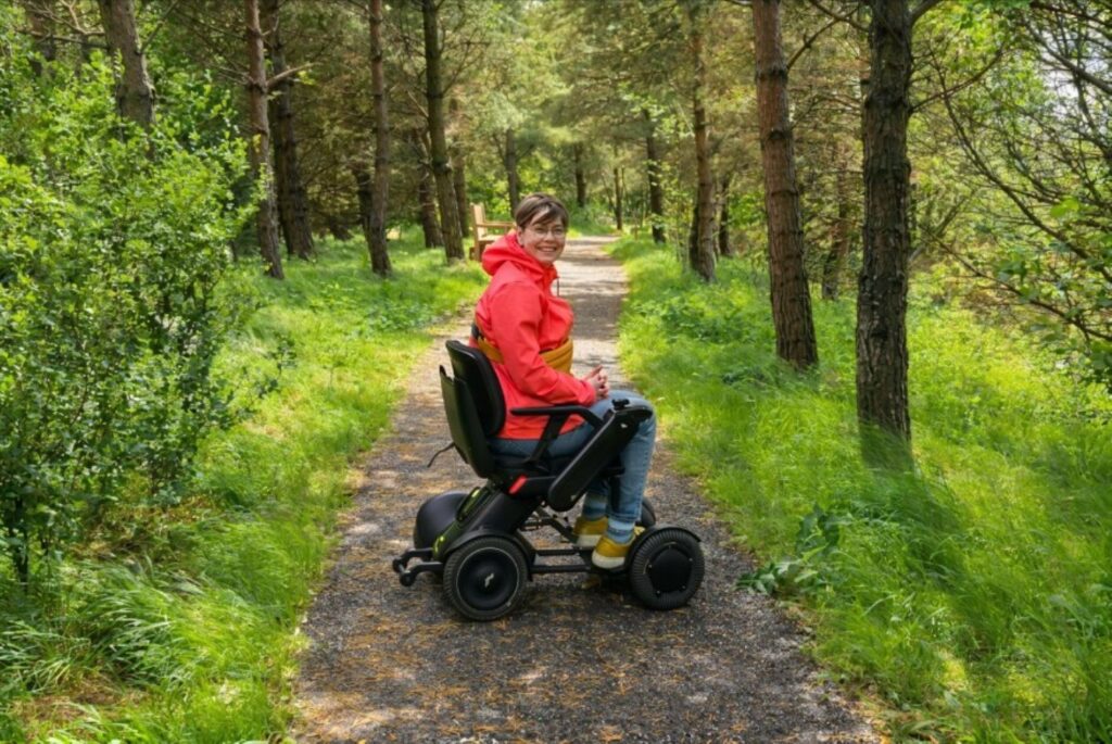 Hadrian's Wall Guide. A powerchair user wearing a bright orange coat smiles a the camera on a track surrounded by lush trees.
