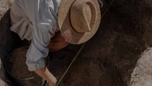 Man crouching over wearing a straw hat in a hole full of soil. Hadrian's Wall Archaeology Forum 2026